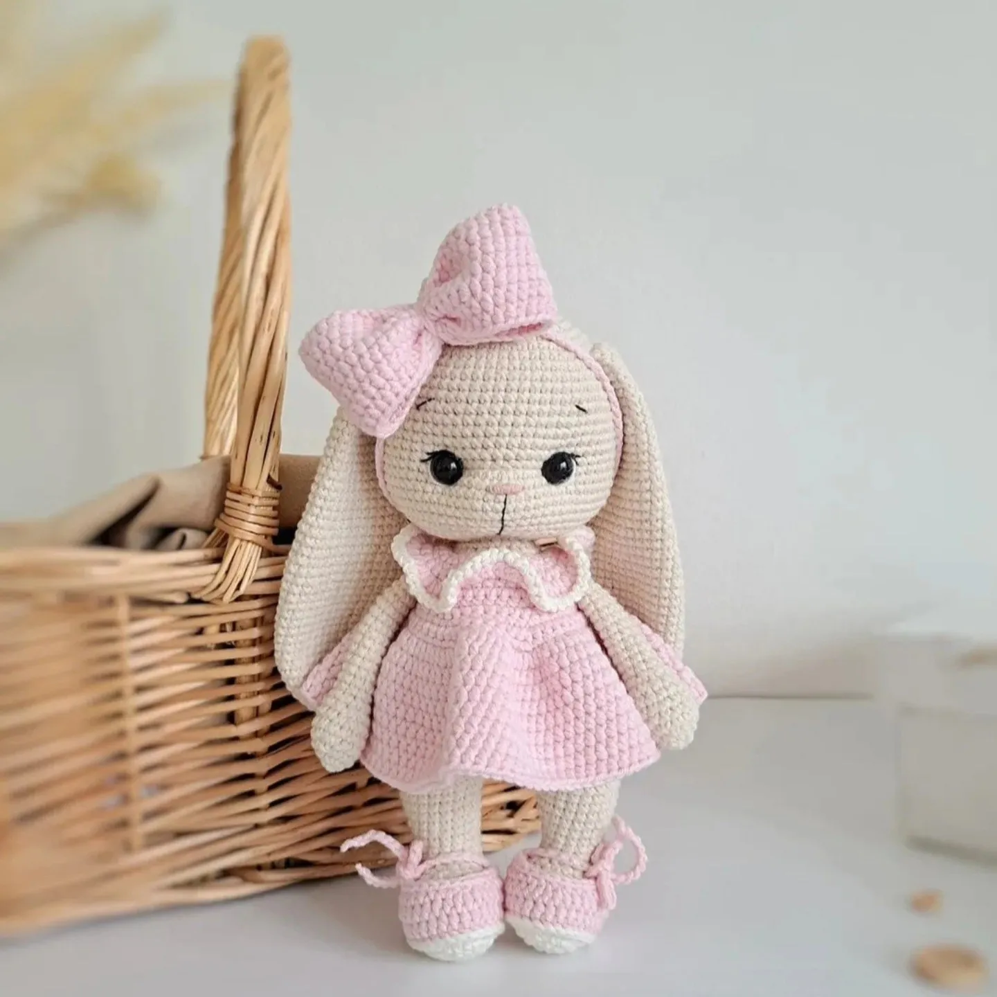 Beige crochet bunny in a pink dress with a bow, standing by a wicker basket on a cream background